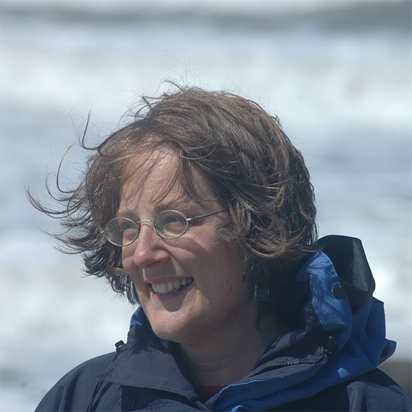 Head and shoulders shot of Susan, looking to the left, wearing a blue waterproof coat and with her hair blowing in the breeze. Foamy white waves are crashing onto the beach in the background.